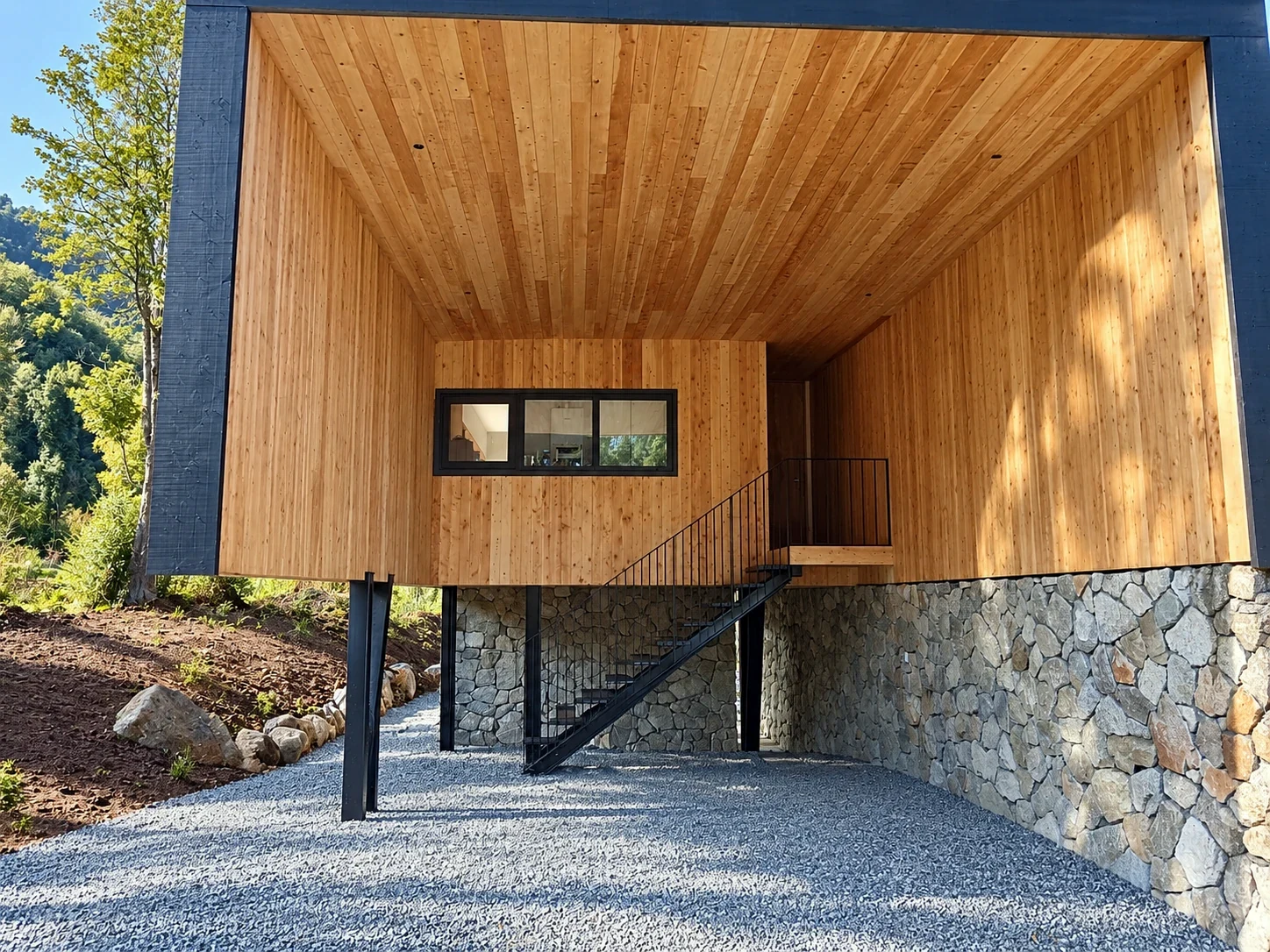Acceso cubierto de vivienda elevada con revestimiento de madera, apoyos metálicos y muro de piedra
